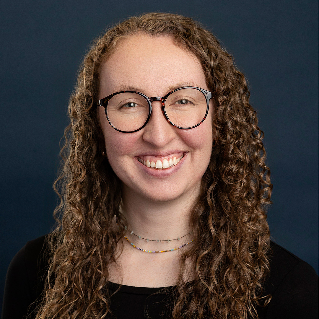 A woman named Makenzie Mathews smiling brightly in front of a gray backdrop bust up.