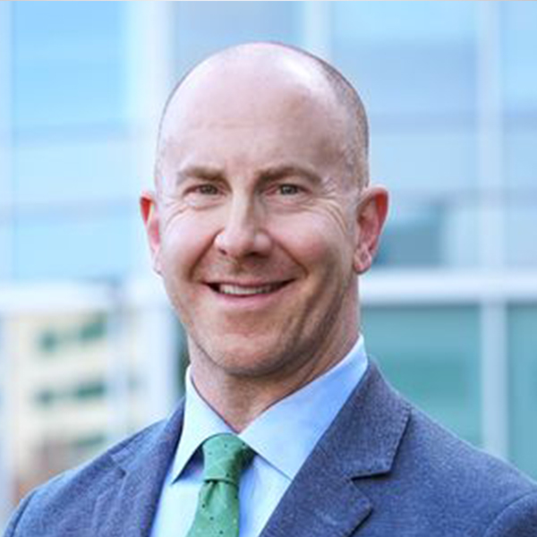 A man named Ryan Fields in a blue suit and green tie in front of a glass building.
