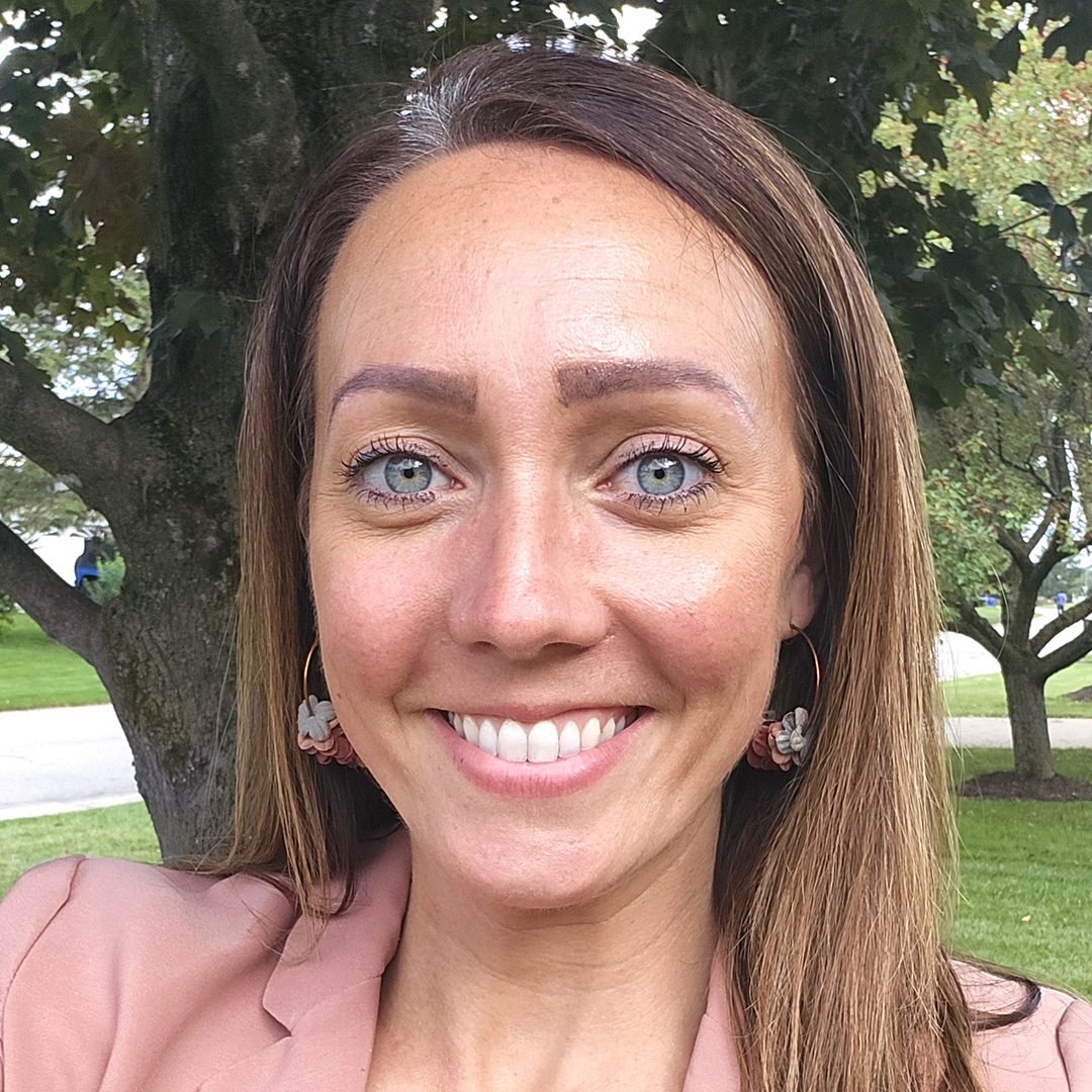 A woman named Caitlin Statt smiling brightly in a pink blazer in from of a large tree.