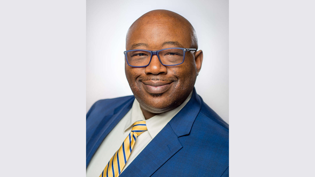 A man named John Blackshear is smiling brightly, wearing glasses and a blue suit.