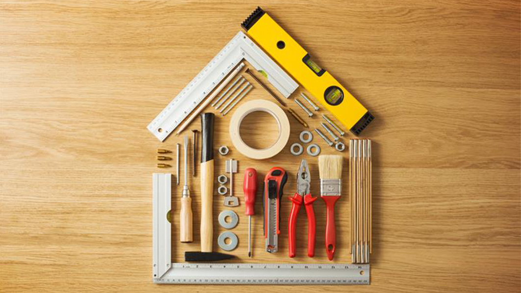 Assorted home repair tools arranged in the shape of a house on a wooden surface, including a level, hammer, pliers, and measuring tools.