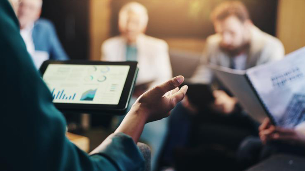 Person presenting data on a tablet during a meeting, with two colleagues listening in the background.