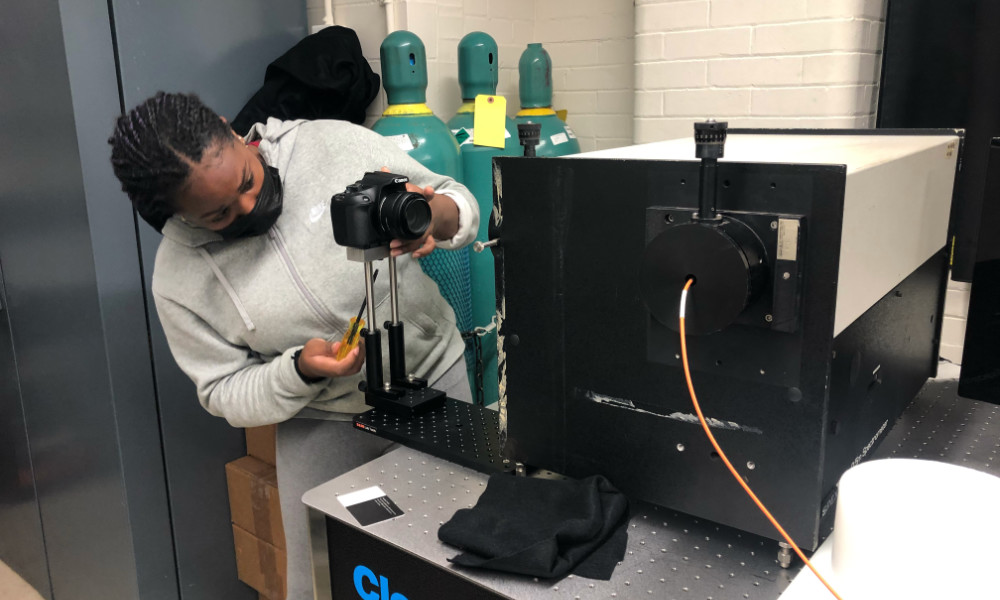 High school student in a mask focuses a camera near a spectrometer.