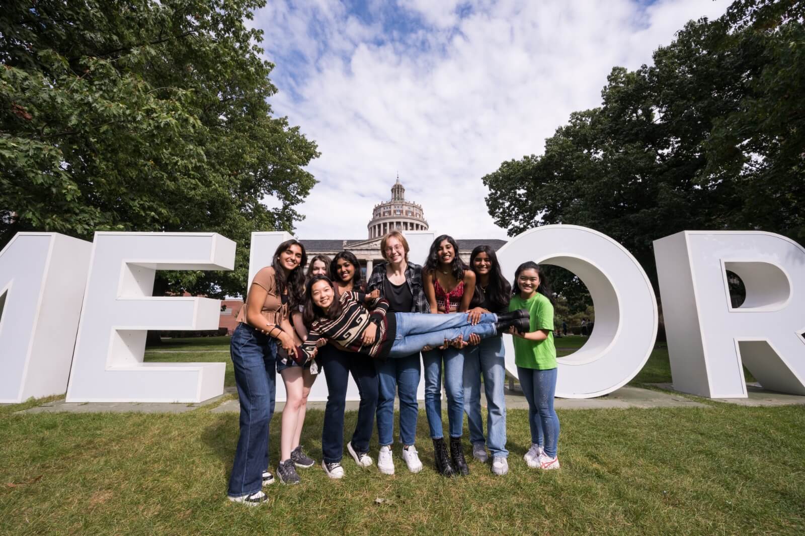 A group of seven young women posing in front of large block letters spelling out "MELIORA." One woman is lifted horizontally by the others. They are standing on grass with trees and a domed building in the background, under a partly cloudy sky.