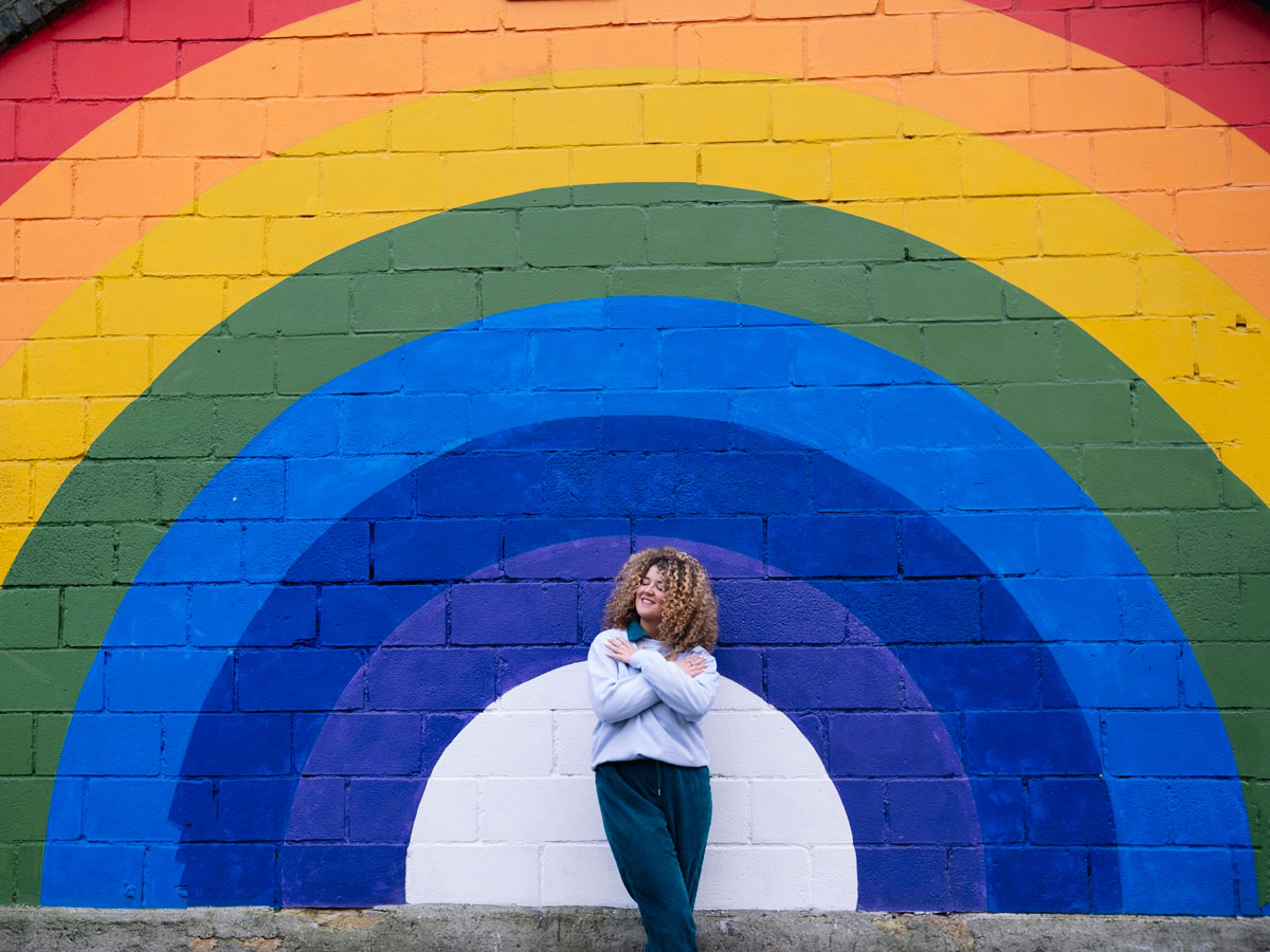 A woman poses confidently in front of a vibrant rainbow wall, showcasing a spectrum of colors behind her.