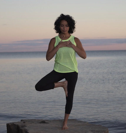 A picture of a female in tree pose with their hands on their heart in front of an ocean.