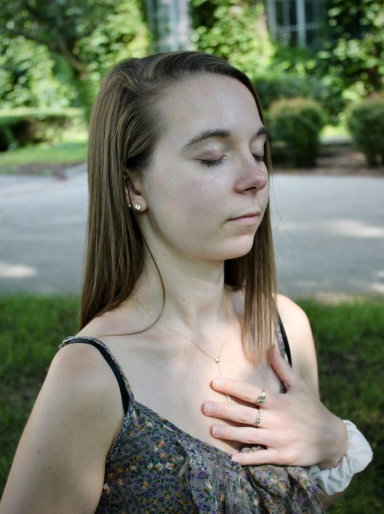 A woman with eyes closed, long brown hair, and left hand on heart in a meditation posture