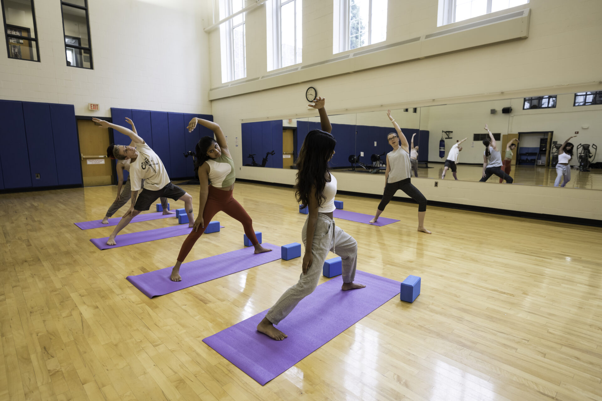 Students practicing yoga facing toward a wall with a mirror, with an instructor at the front of the room