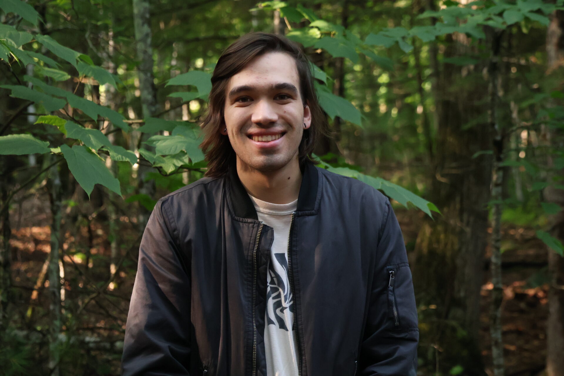 A young male smiling with a tree behind him, with long dark hair and a leather jacket on.