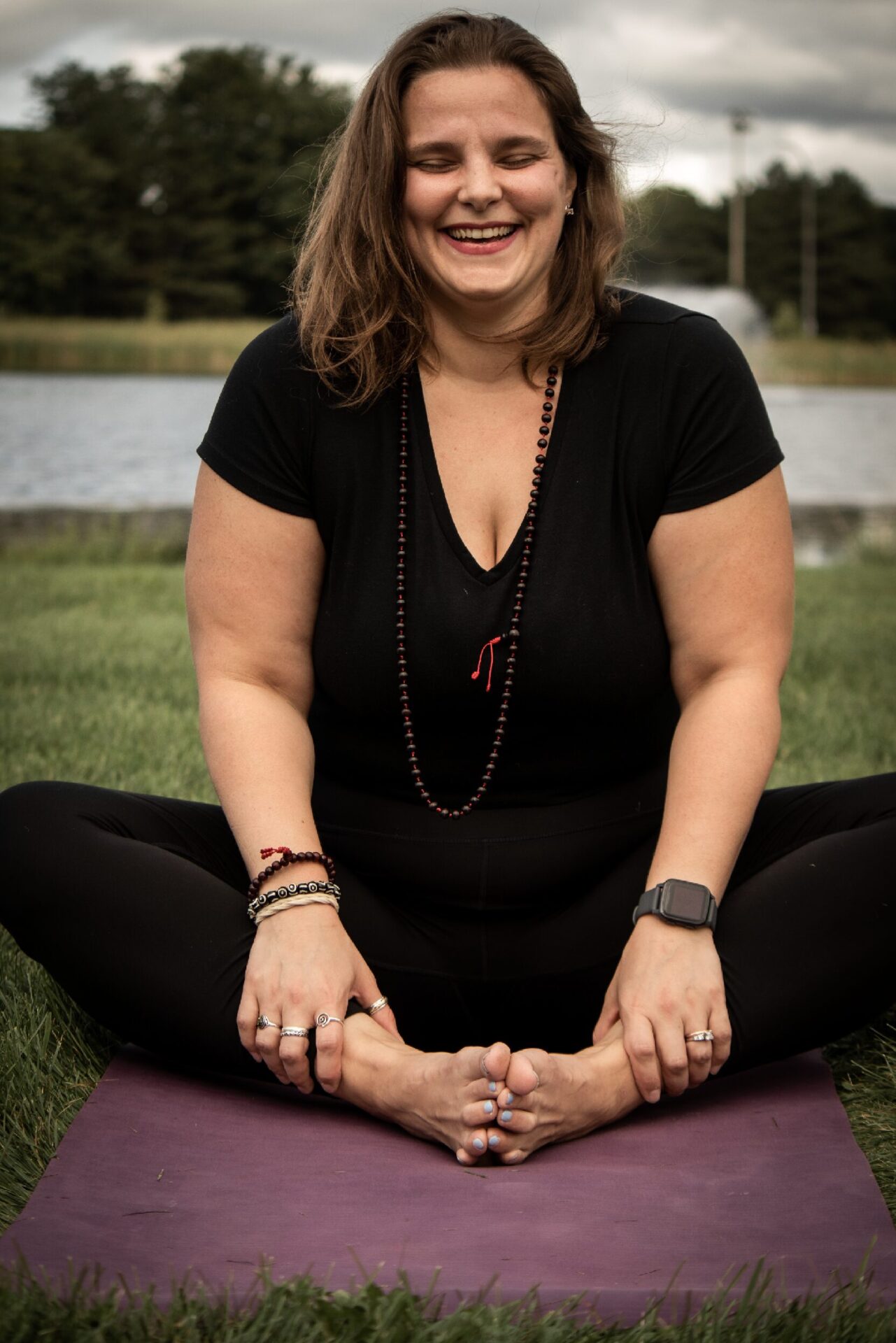 A woman sitting on a yoga mat outside with a black shirt and pants on in a seated pose smiling