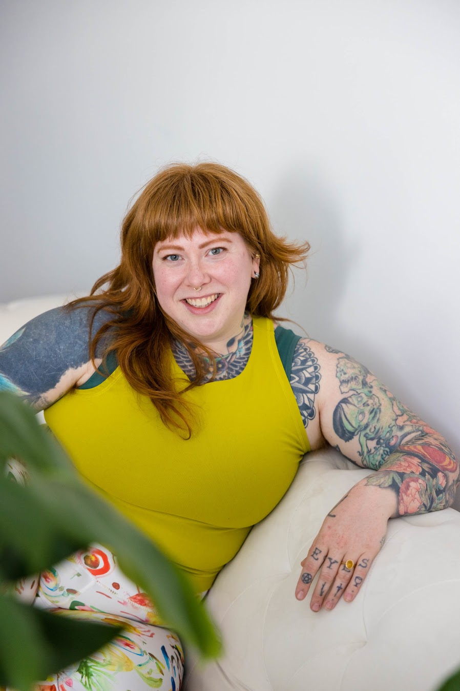 An individual in front of a white wall with athletic clothing on, tattoos on their arms, and long, orange hair.