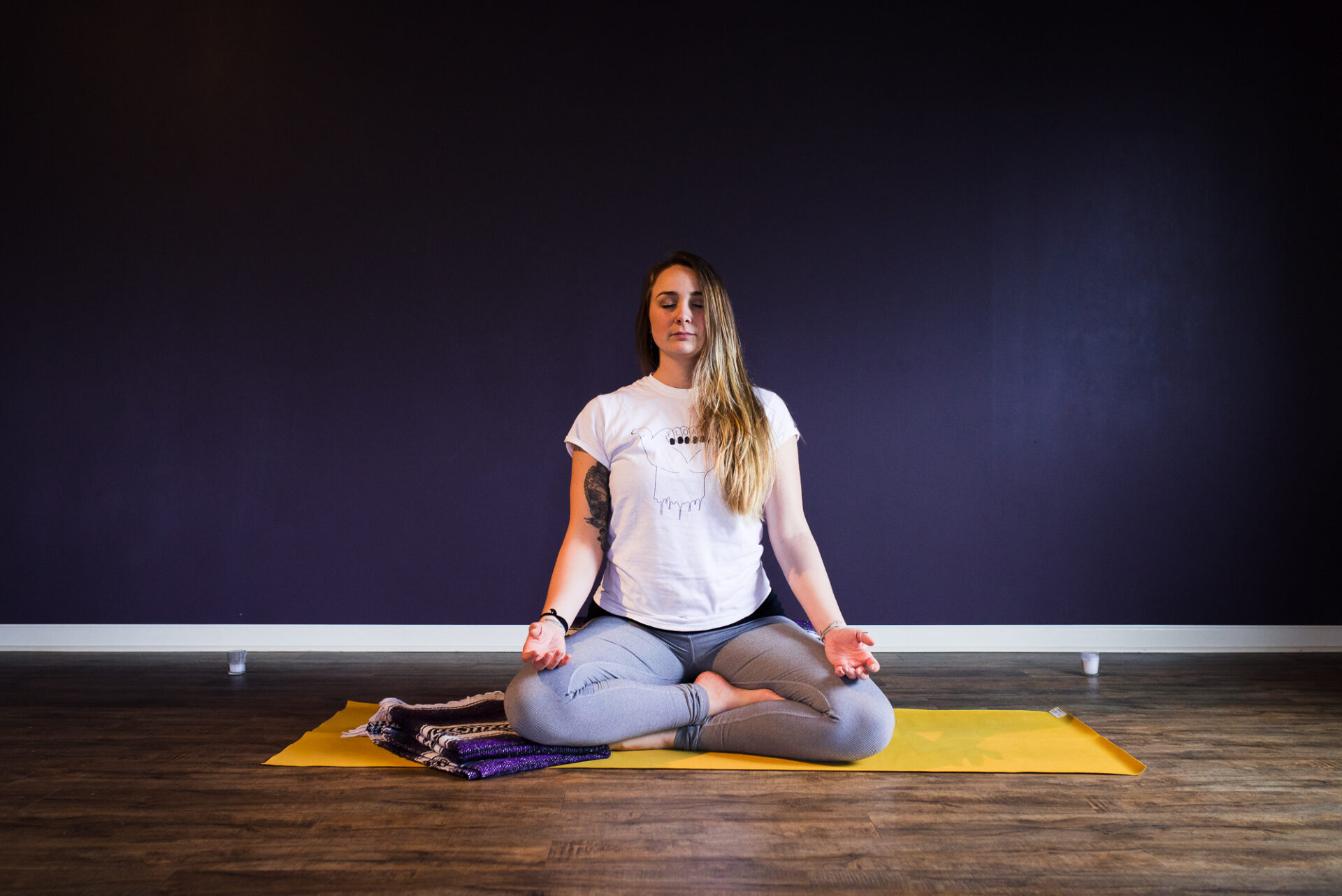 A woman on a yoga mat in athletic clothing in front of a purple wall in a meditation posture