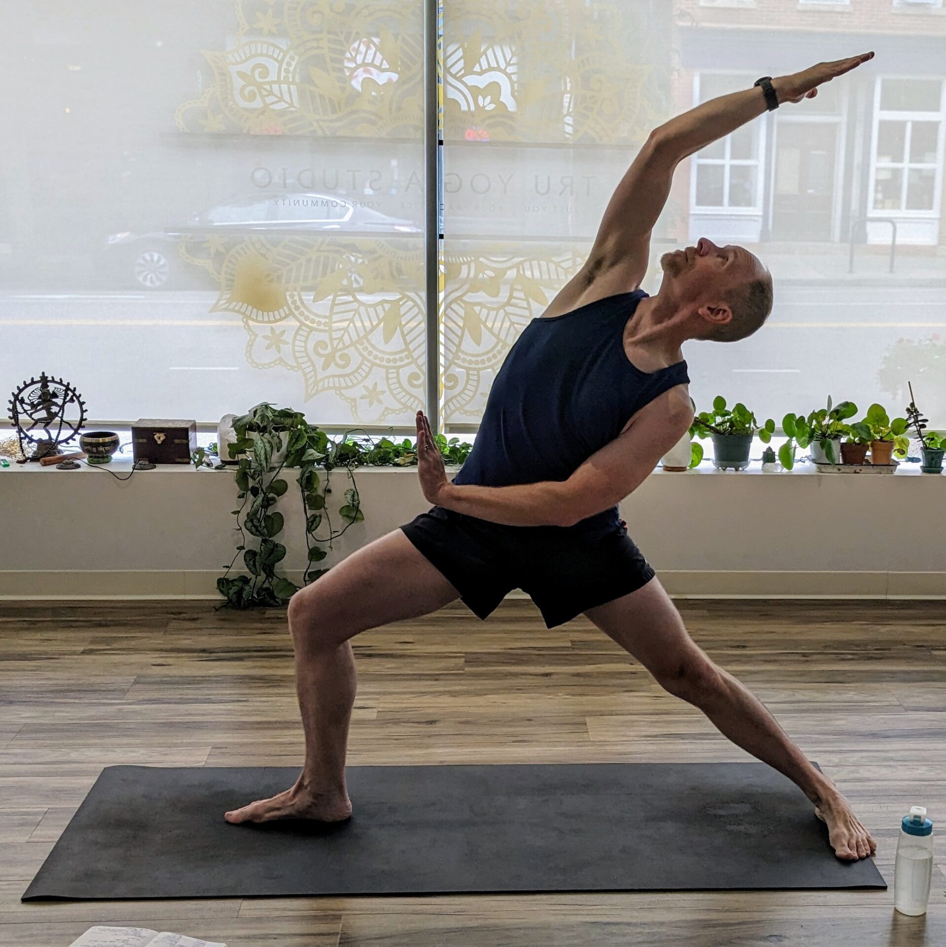A photo of a man on a yoga mat wearing black tanktop and shorts in peaceful warrior yoga pose