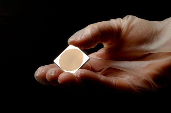 gloved hand holds a small square of gold-colored metal.
