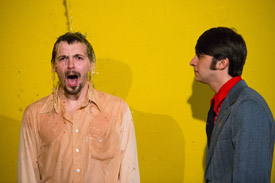 Two actors in front of a yellow backdrop being splashed with water.