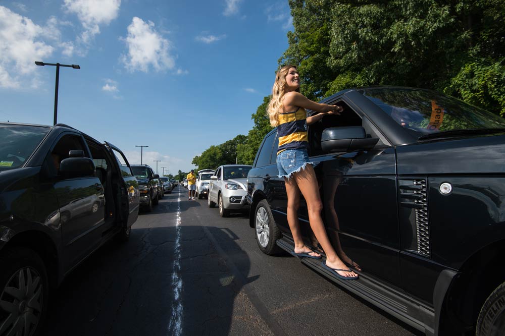 long line of cars in a parking lot, with one student hanging off the side of one