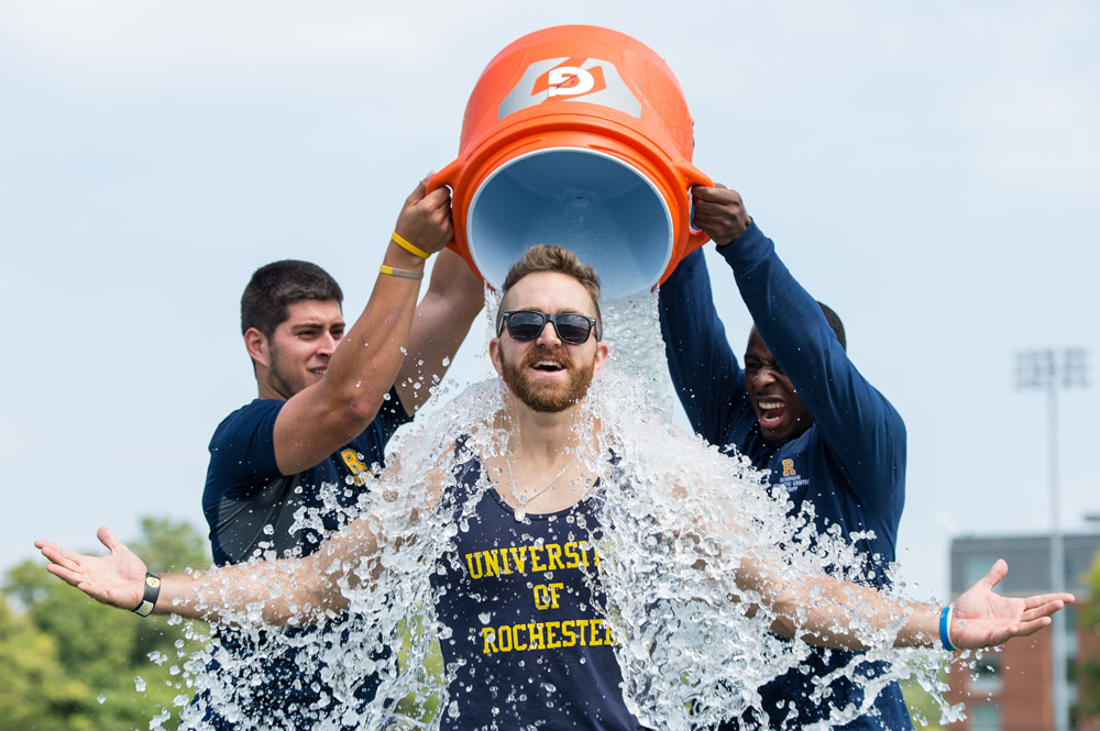 Feature image coach wearing University of Rochester uniform smiles as football players dump large bucket of water over his head