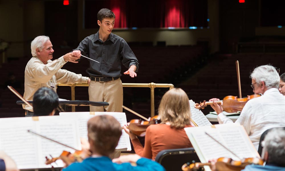Feature image instructor holds the baton arm of a young conducting student who stands at the podium in from of orchestral musicians