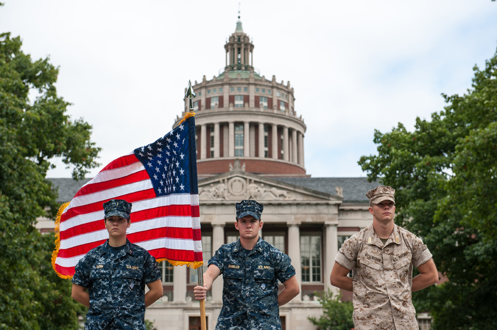 Feature image three NROTC midshipmen in uniform in front of Rush Rhees Library, one of them holding an American flag