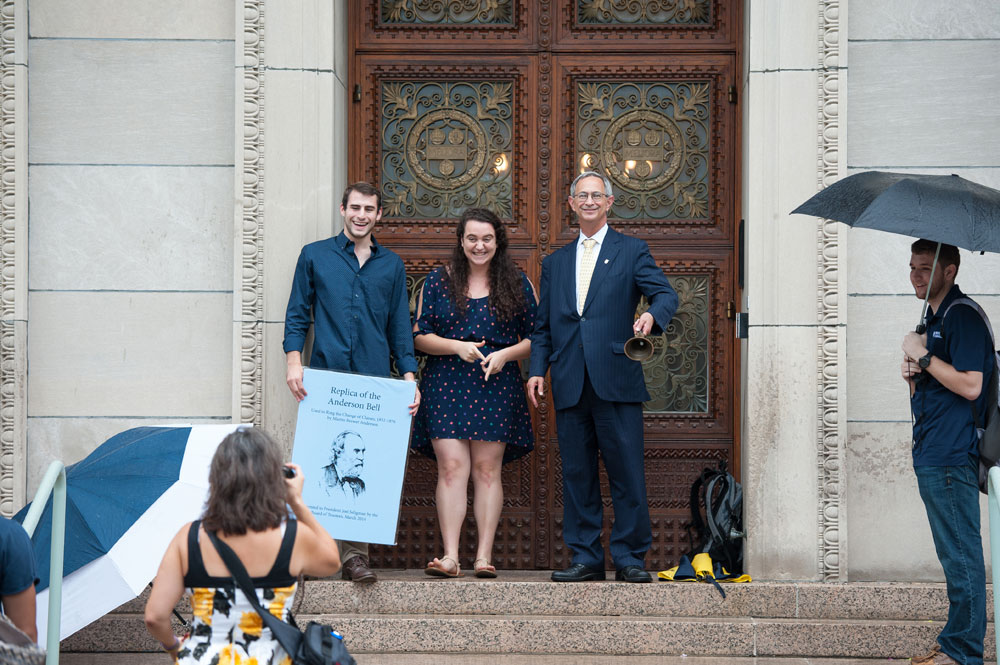 Feature image two students with President Seligman on the steps of the library, as Seligman rings a handheld bell