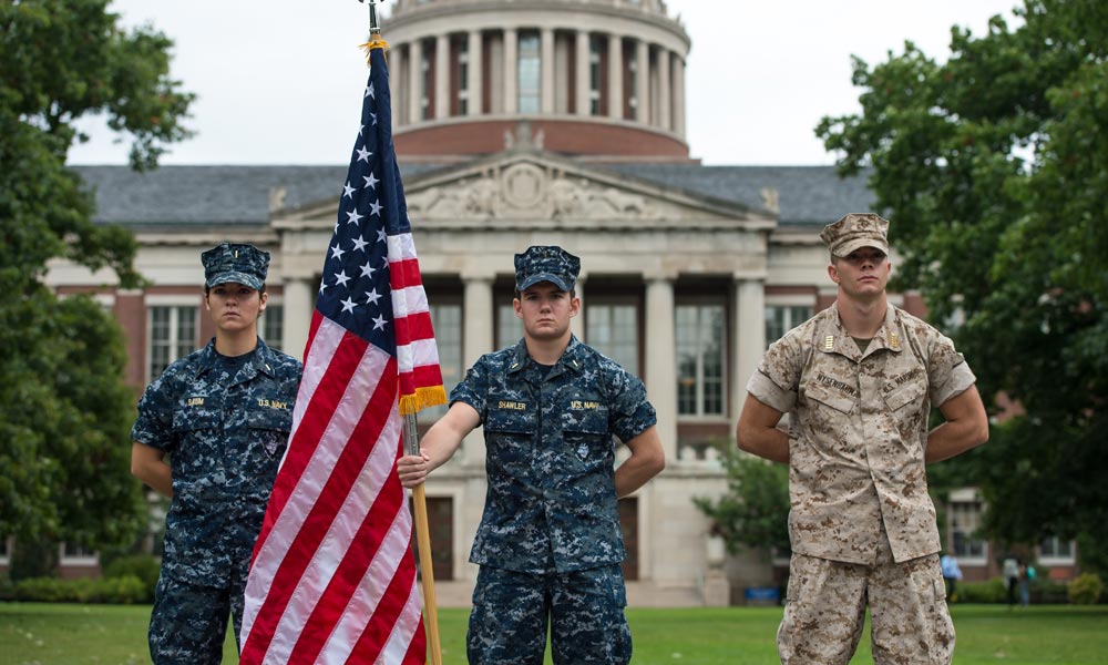 three students in military fatigues, one holding an American flag, standing in front of Rush Rhees Library