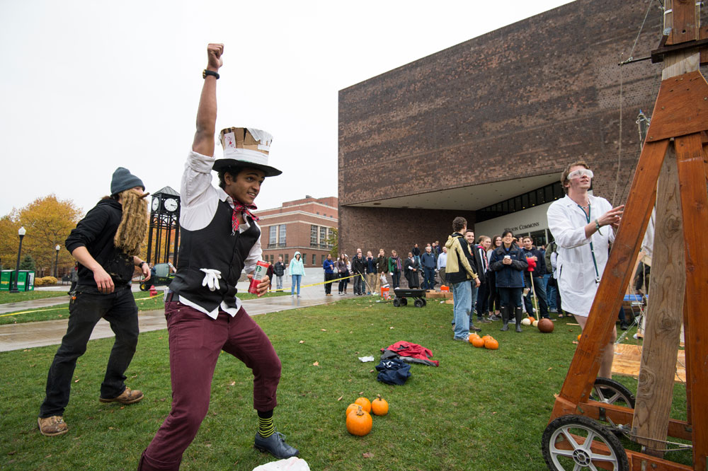 Feature image student standing next to a trebuchet and a pile of pumpkins pumps his fist in the air