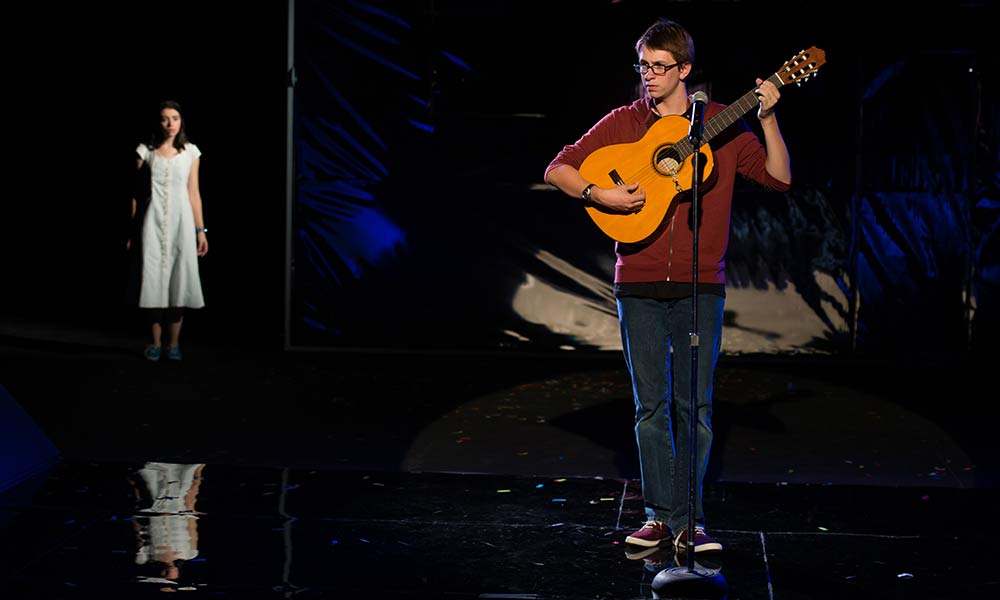 Feature image young man playing guitar on stage with woman in white behind