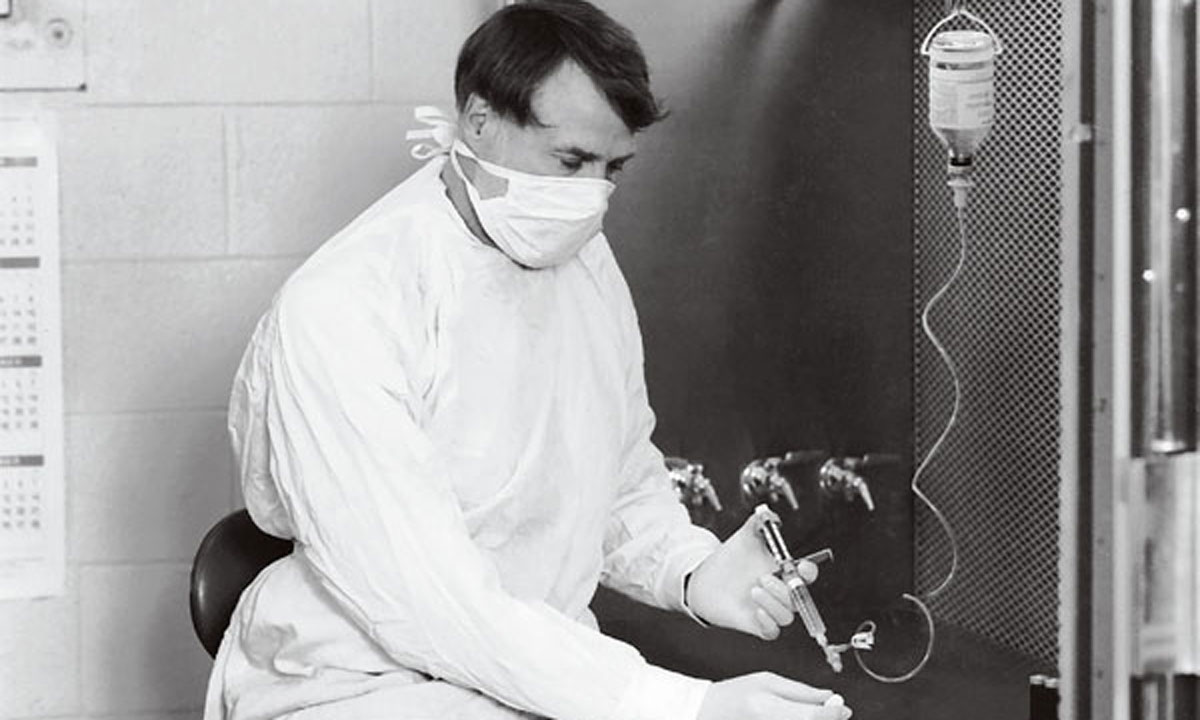A man in a white coat and surgical mask sits by a machine at the University of Rochester, focusing on Hib infections research.