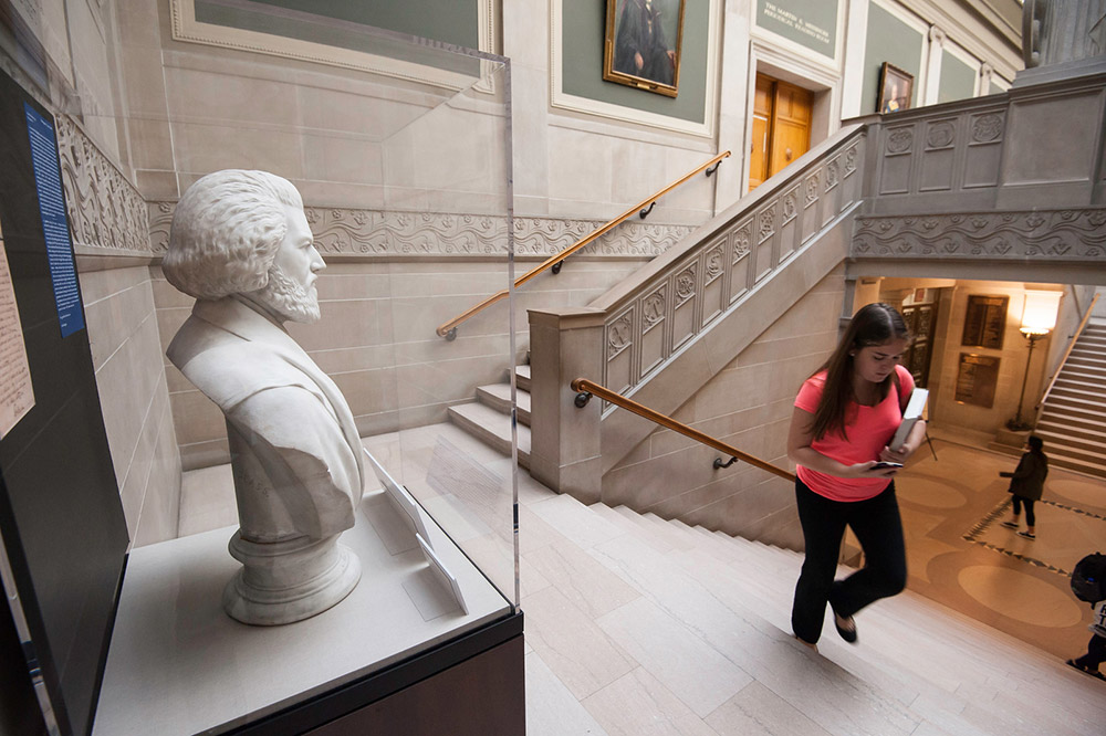 Feature image statue of Frederick Douglass in a glass case on the library staircase as people walk past