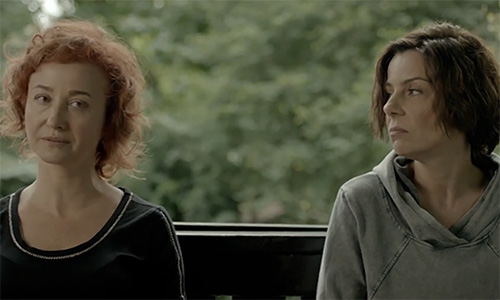Feature image still from film shows two women sitting at a table, with angry expressions on their faces