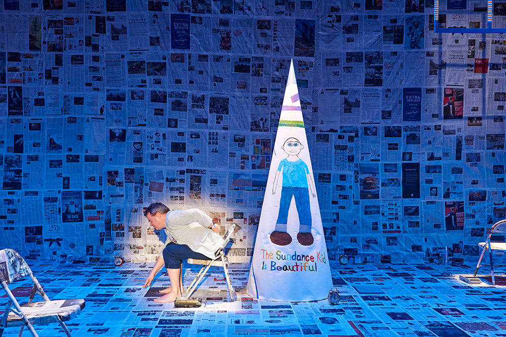 Feature image man sits in the spotlight on a stage covered in newspapers