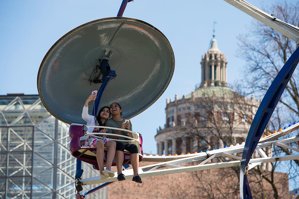 students take photos on a carnival ride, with Rush Rhees tower in the background