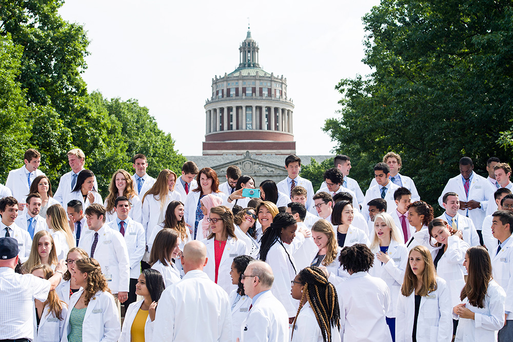Feature image students in white doctor coats pose for photos in front of Rush Rhees Library