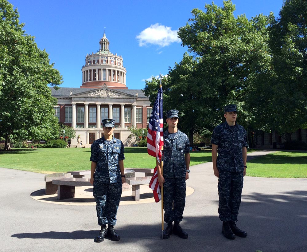 three midshipmen stand at attention on the quad, with Rush Rhees Library in the background