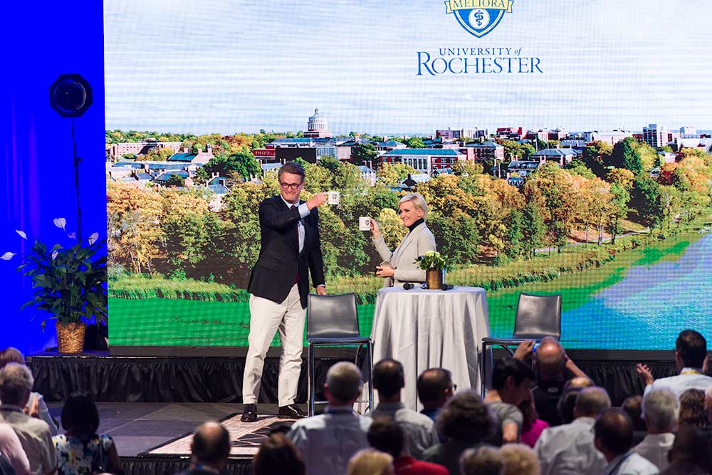 Joe Scarborough and Mika Brzezinski on stage holding University of Rochester coffee mugs