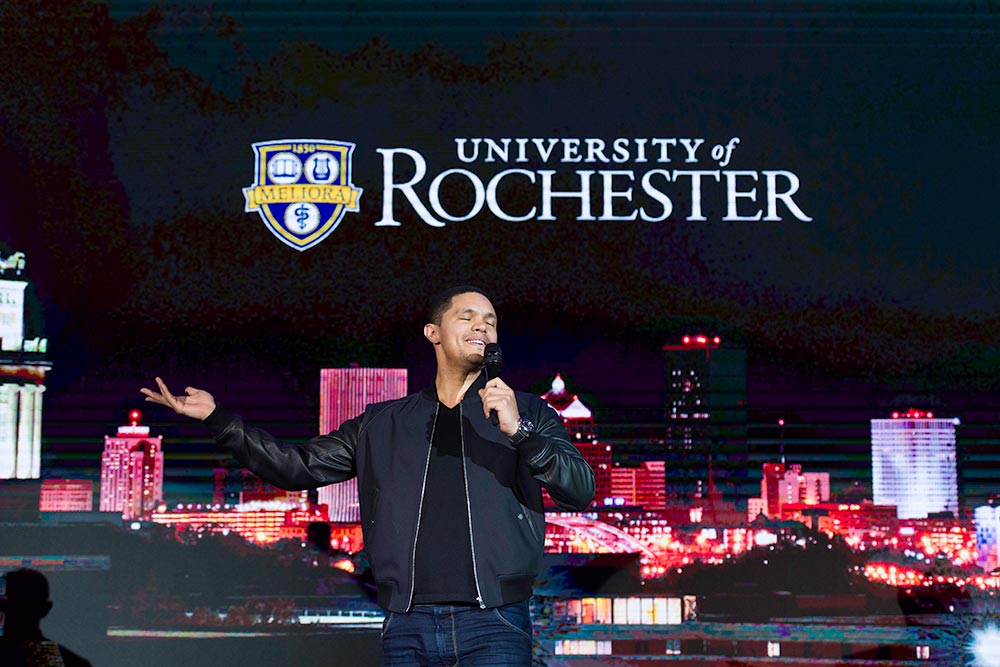 Trevor Noah on stage in front of a University of Rochester logo. 