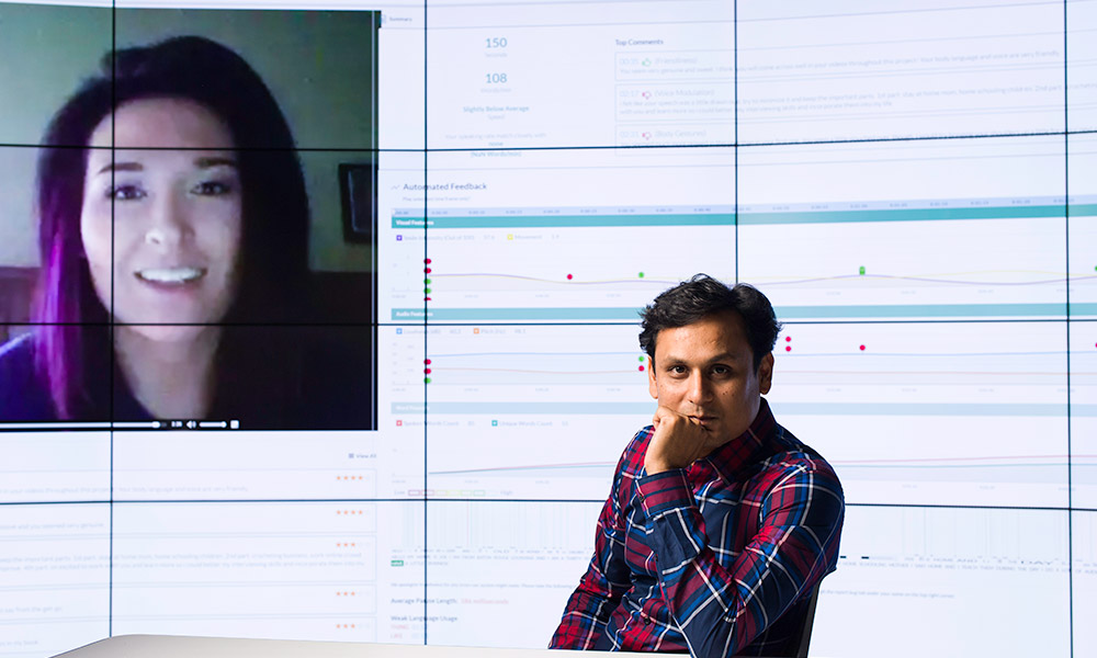 researchers sits in front of a large screen that contains the image for a human face, speaking