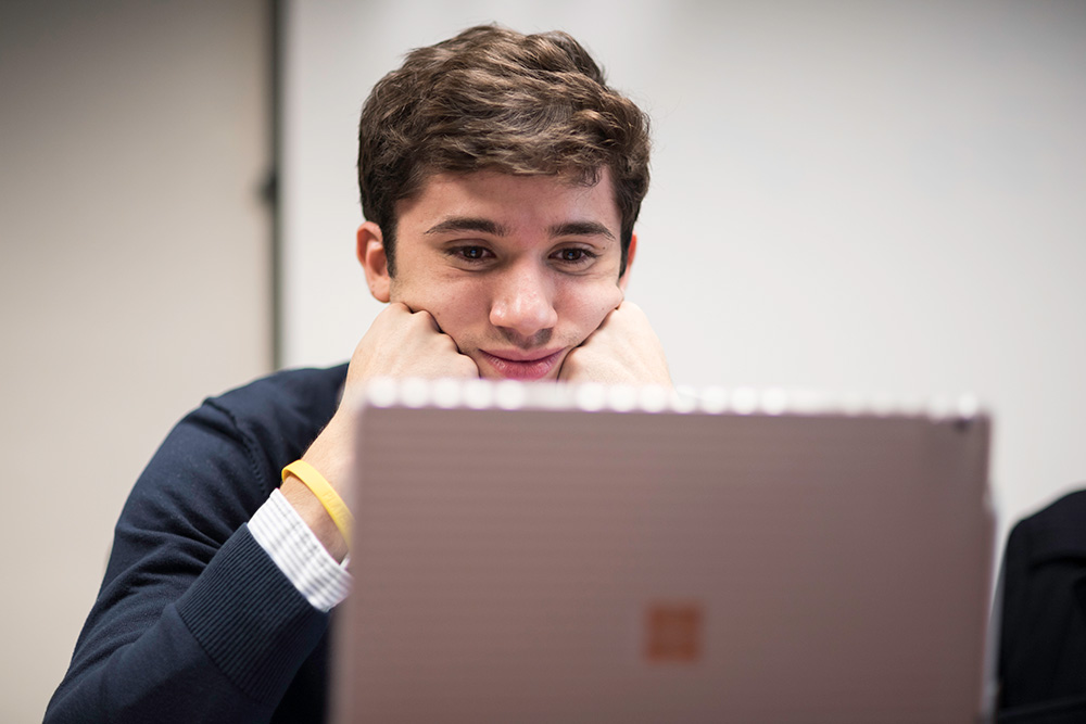 Feature image student sits behind a laptop screen, smiling with his chin in his hands