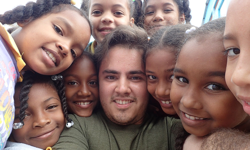 Feature image student takes a selfie, smiling, surrounded by smiling kids