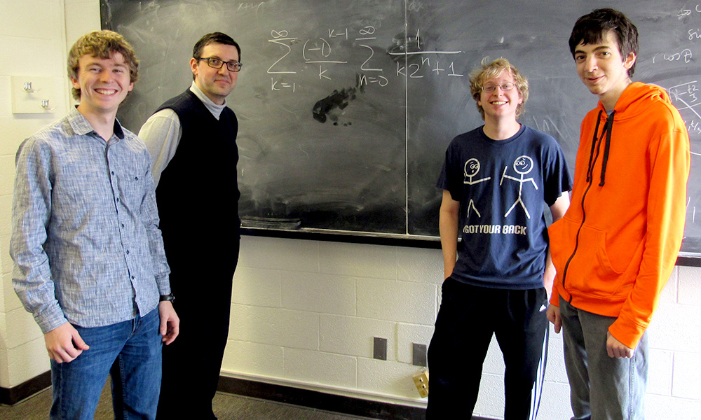 three students and their professor pose in front of a blackboard with complicated math problems on it