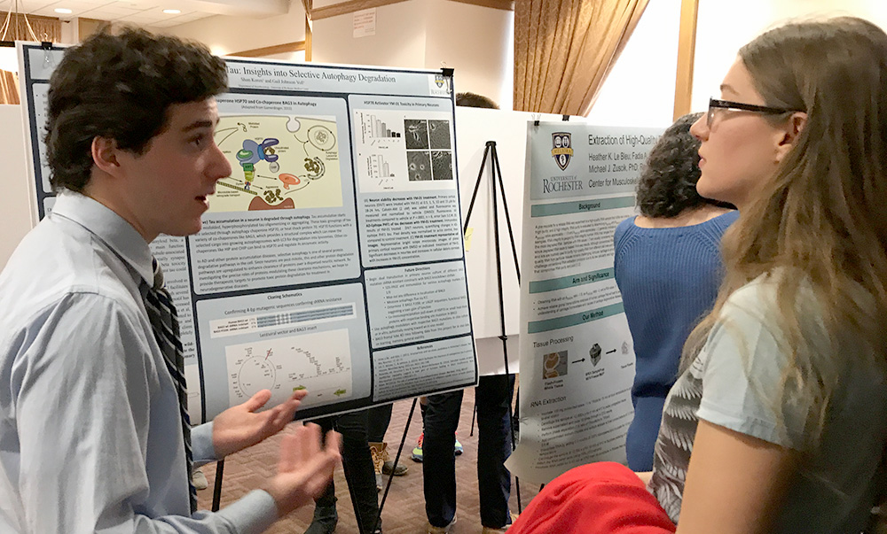 student stands in front of poster, speaking about his research to another student