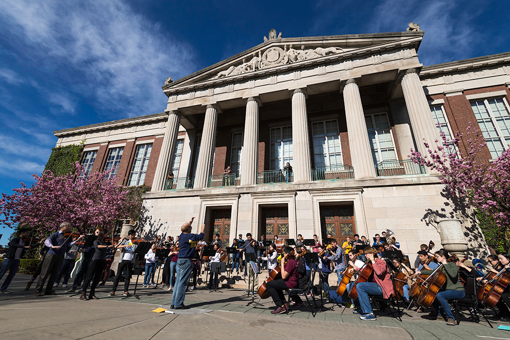 Feature image conductor leads an orchestra of musicians in front of Rush Rhees Library