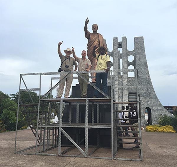 three men stand on a scaffold under a statue, pointing in the same manner as the statue