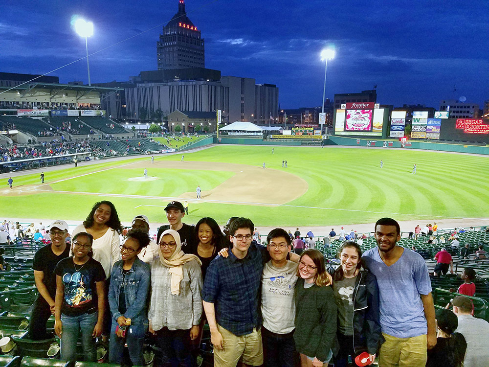 Feature image group of students pose for a photo a Frontier Field, with Kodak tower and Rochester skyline in background