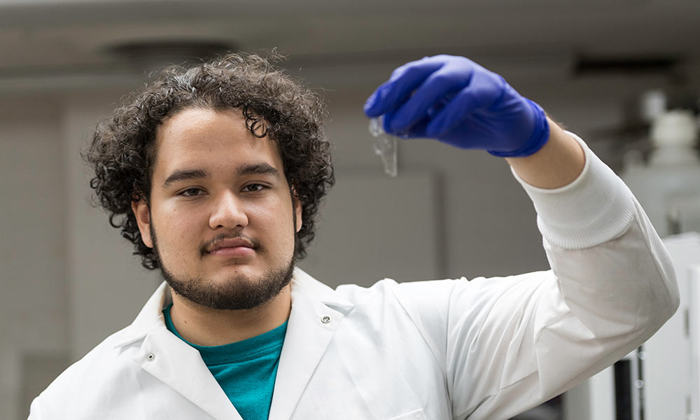 young man in a lab coat, holding up a transparent polymer in a gloved hand