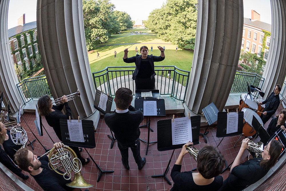 conductor raises arms and faces musicians playing on a balcony