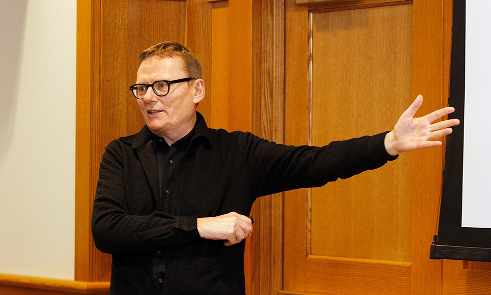 man gestures while speaking in front of audience in library