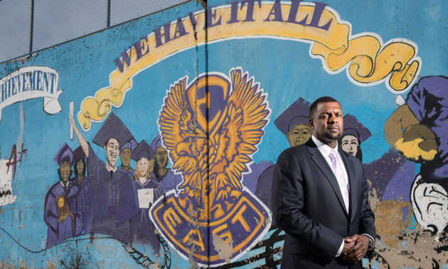 Shaun Nelms stands in front of a painted mural that reads WE HAVE IT ALL : EAST HIGH