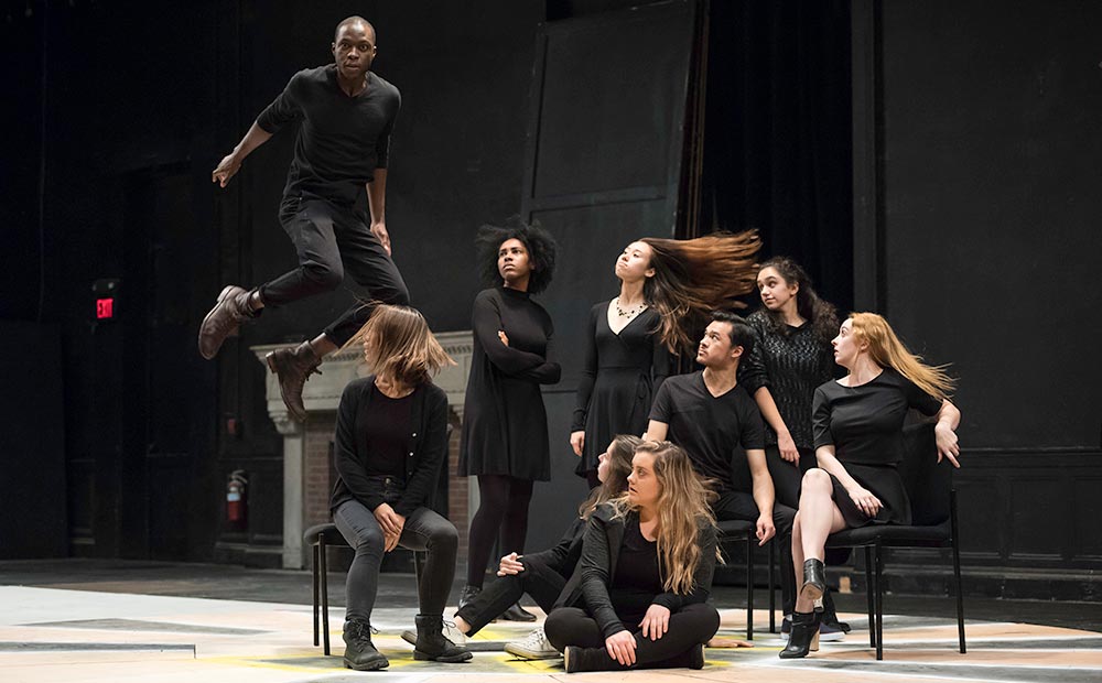 Feature image cast of actors on stage, one of them leaping through the air.