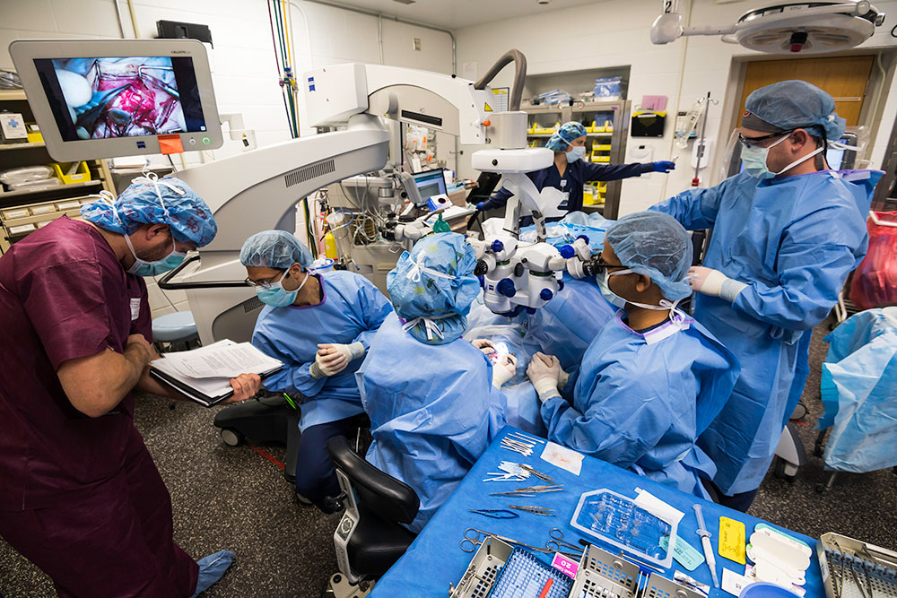 large group of surgeons in operating room, with surgery visible on small computer monitor